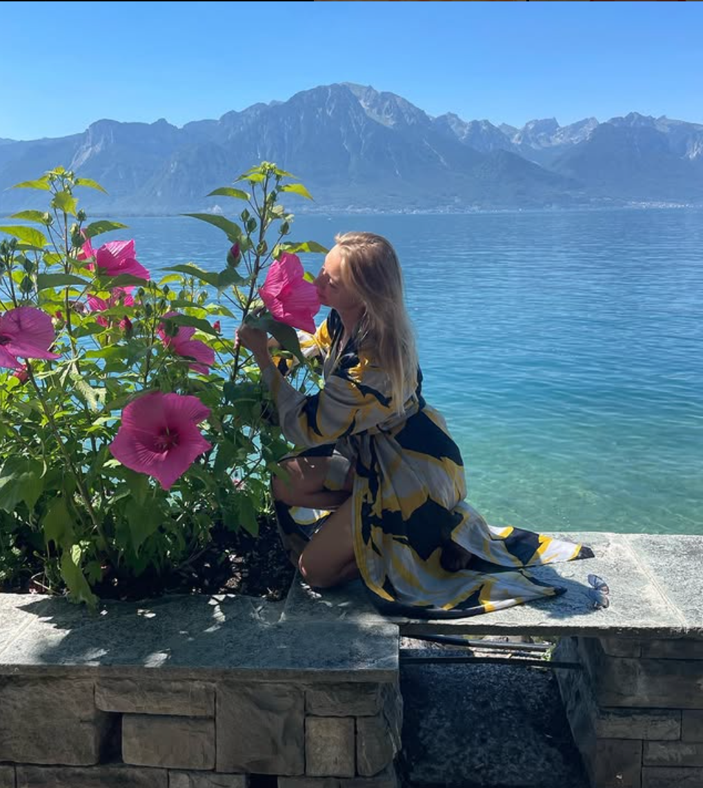 Olesia Ra kneeling beside pink hibiscus flowers in front of a mountain lake, wearing one of her first kimono designs from Rai Kimonos.