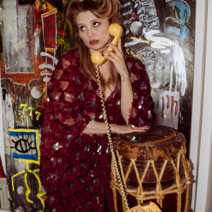 A woman in a textured red outfit poses with a vintage yellow telephone, surrounded by colorful abstract art and a tribal drum in an indoor setting.